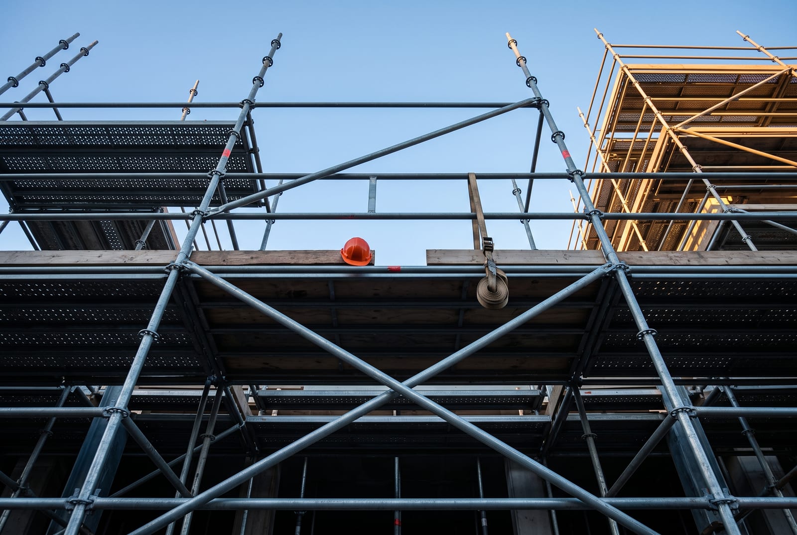 Empty scaffolding at dawn seen from the ground, a section of guardrail missing on the third lift, a ratchet strap hanging loose and a hard hat left on the planking