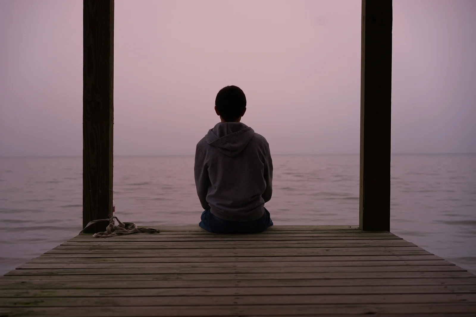 A hooded young person seated alone on a wooden dock at sunset, contemplative, family-recognizable, the calm visual the parent of a struggling teenager is looking for