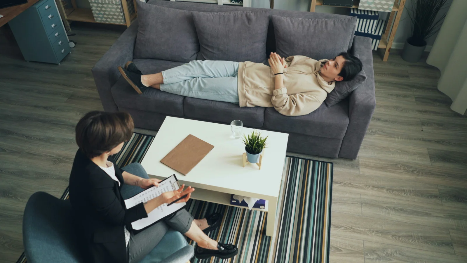 Top-down view of a clinician with a clipboard and a patient lying on a couch in a calm consult room, demystifying what happens at a first psychiatric visit