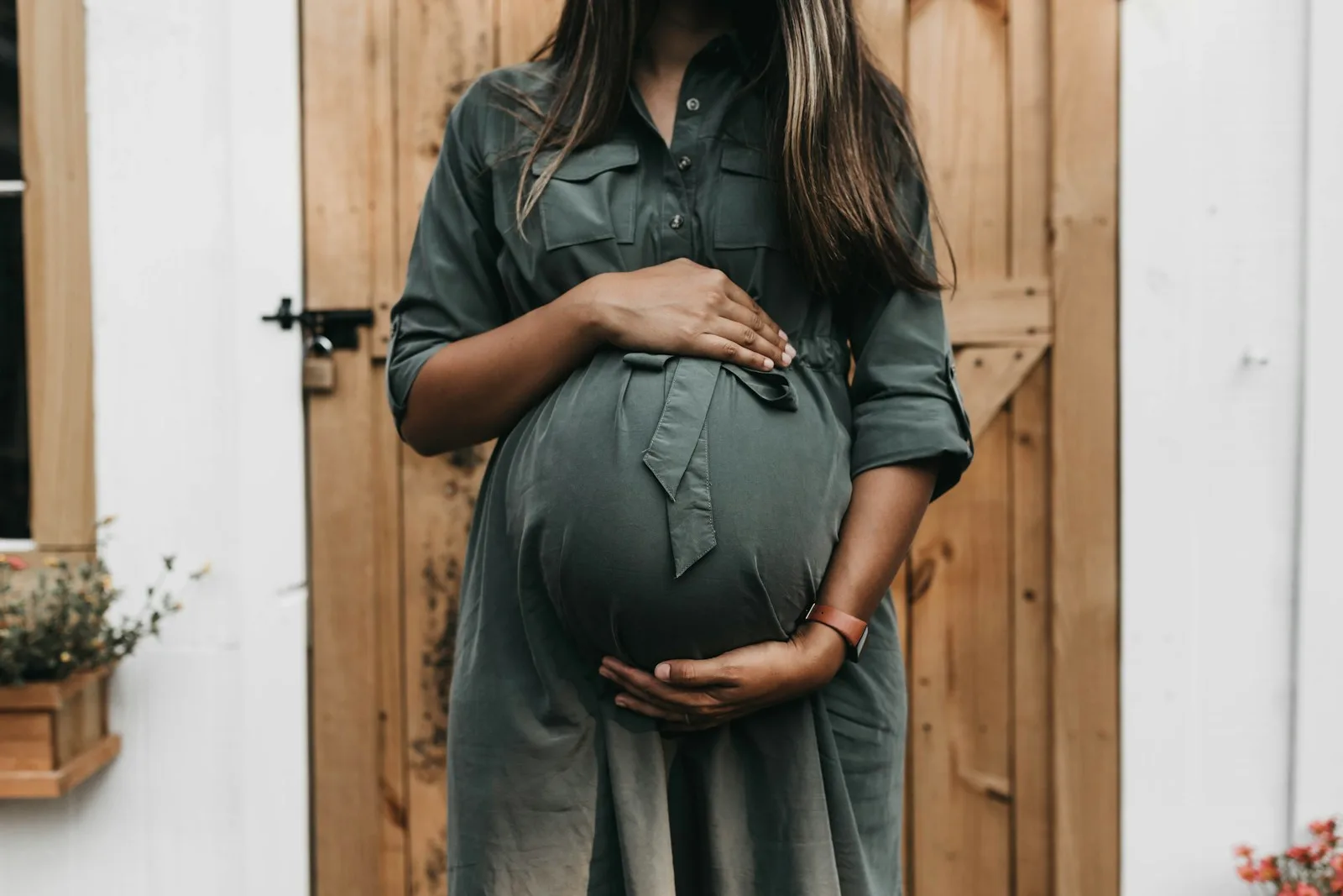 A pregnant woman in a sage green shirt dress rests both hands on her belly in warm light, the patient this page plans treatment with rather than around