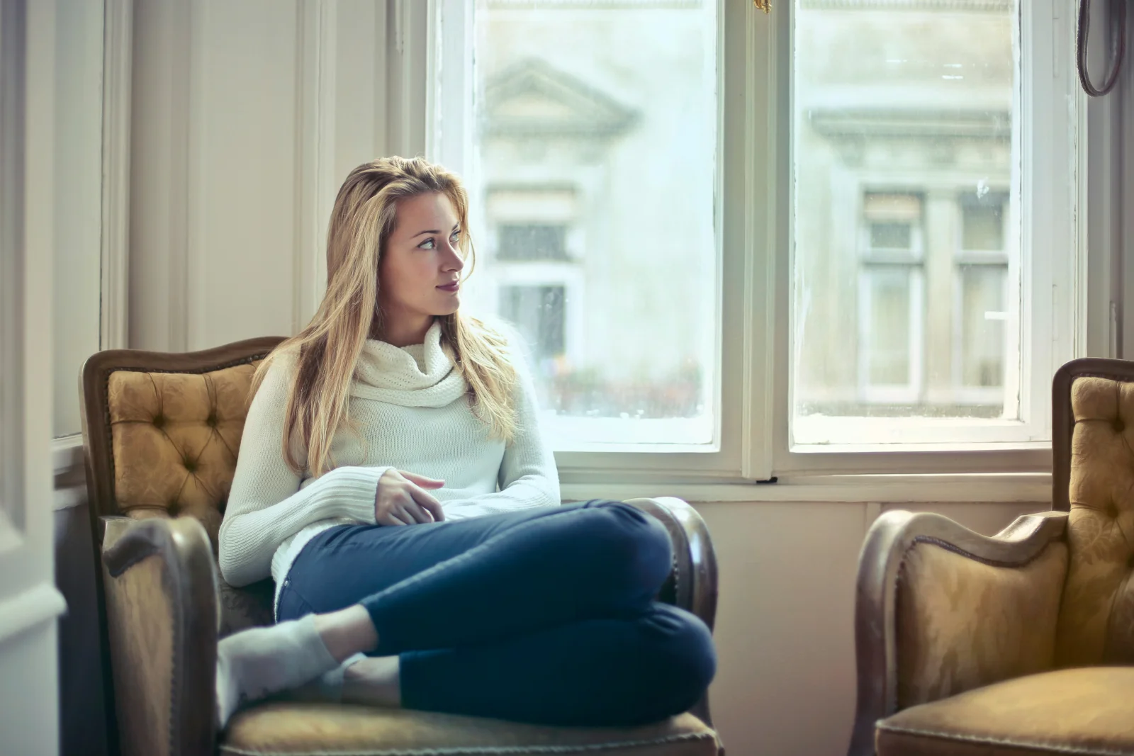 A patient seated in a tufted chair by a tall window, contemplative, capturing the moment of reaching out at the end of a long day
