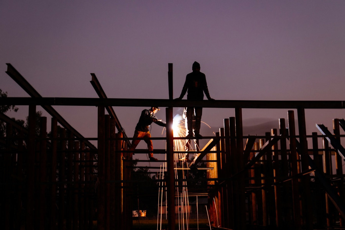 Two ironworkers on structural steel at dusk, one welding, sparks falling through the frame