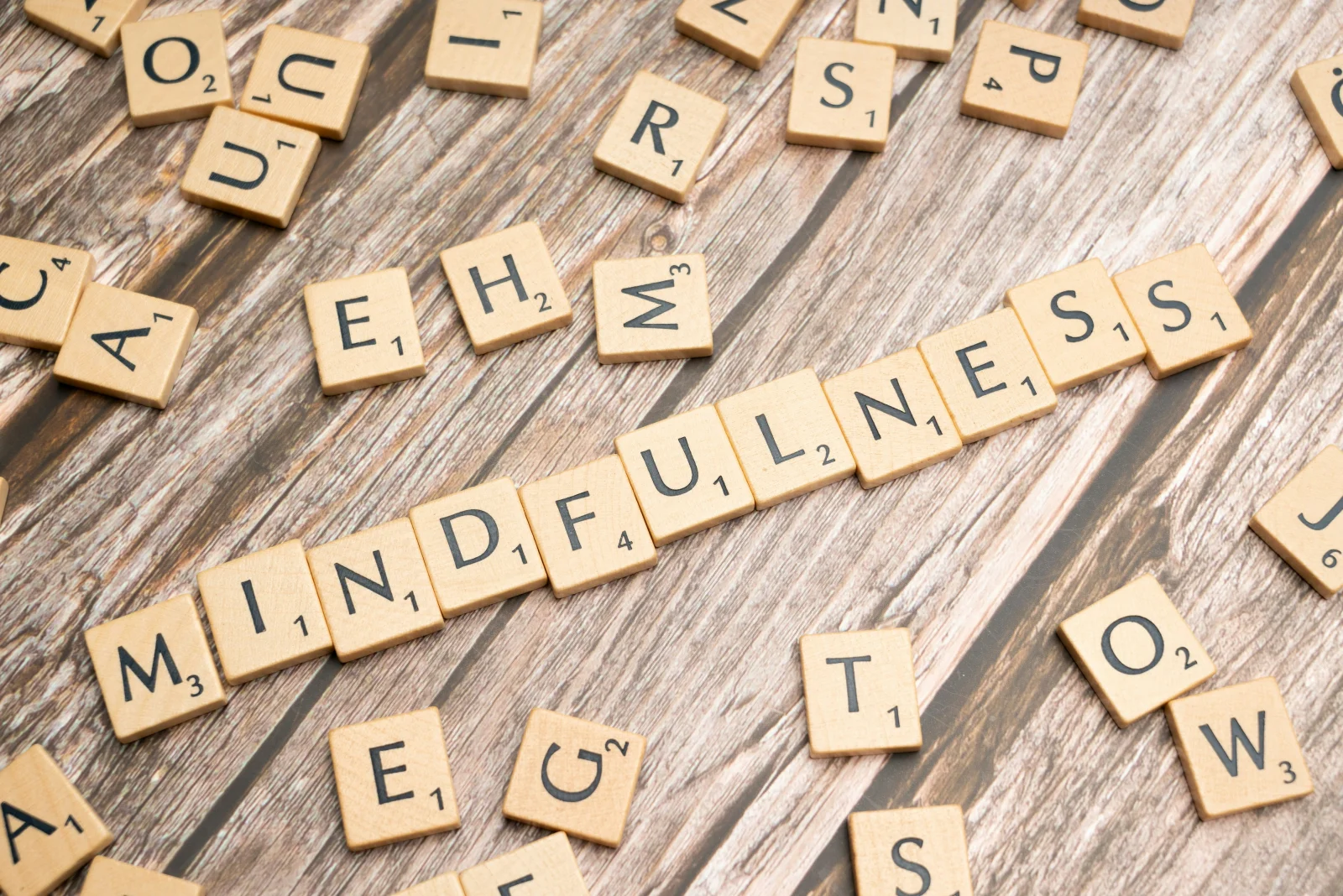 Scrabble tiles spelling MINDFULNESS on a weathered wood surface, the practice's clinical philosophy rendered as a quiet header motif rather than a smiling-staff cliche